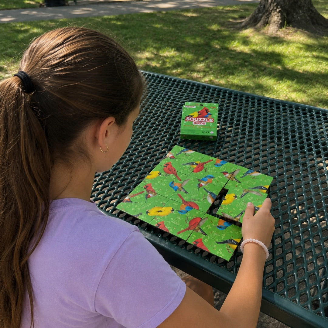A person arranging nine Birds Squzzle picture tiles at an outdoor table, with the matching box nearby.